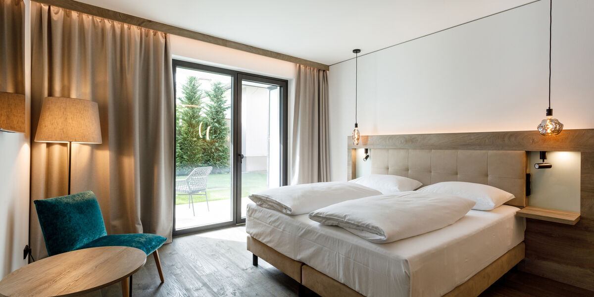 Modern hotel room with double bed, wooden accents, teal-colored chair and large window overlooking a garden.