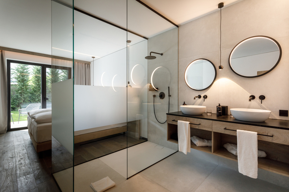 Modern bathroom with double washbasin, round mirrors, glass shower and a bedroom visible through a glass wall.