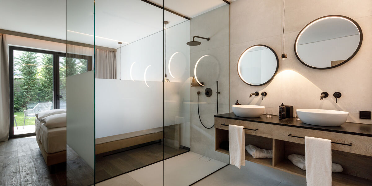 Modern bathroom with double washbasin, round mirrors, glass shower and a bedroom visible through a glass wall.