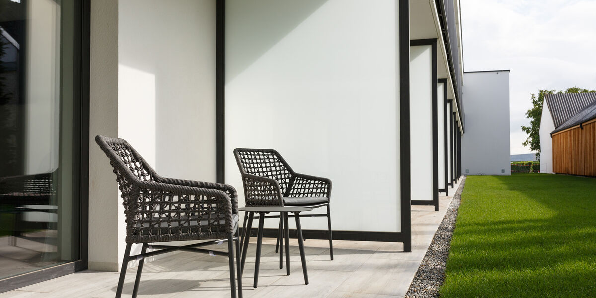 Two black woven chairs and a small table on a modern terrace next to a grassy lawn and a white building.