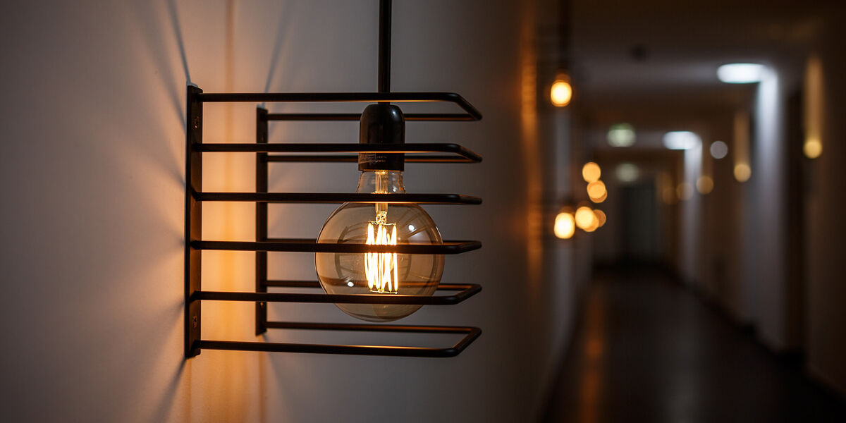 A close-up of a glowing light bulb in a modern black metal wall light lining a dimly lit hallway.