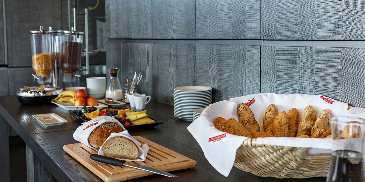 Breakfast buffet with cold cuts, breadsticks, fruit, cereal dispensers and plates on a dark counter.
