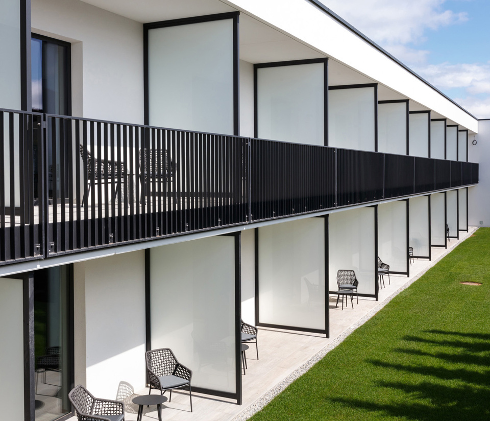 Modern building with large frosted glass windows, black railings and outdoor chairs on terraces at the edge of a green lawn.