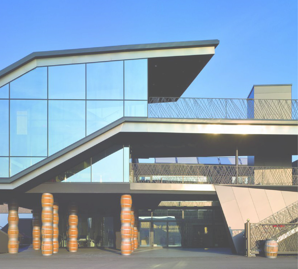 Modern building with angular glass façade, external staircase and stacked barrels near the entrance under a blue sky.