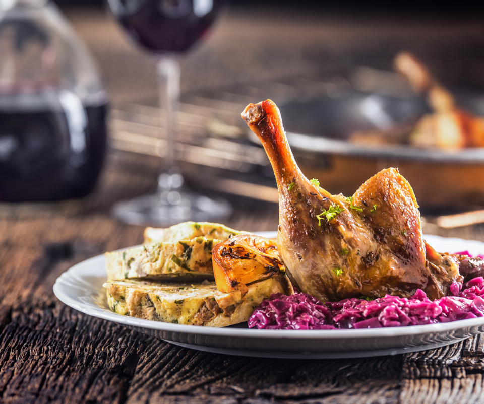 Roast leg of duck with bread dumplings and red cabbage on a white plate, with wine in the background.